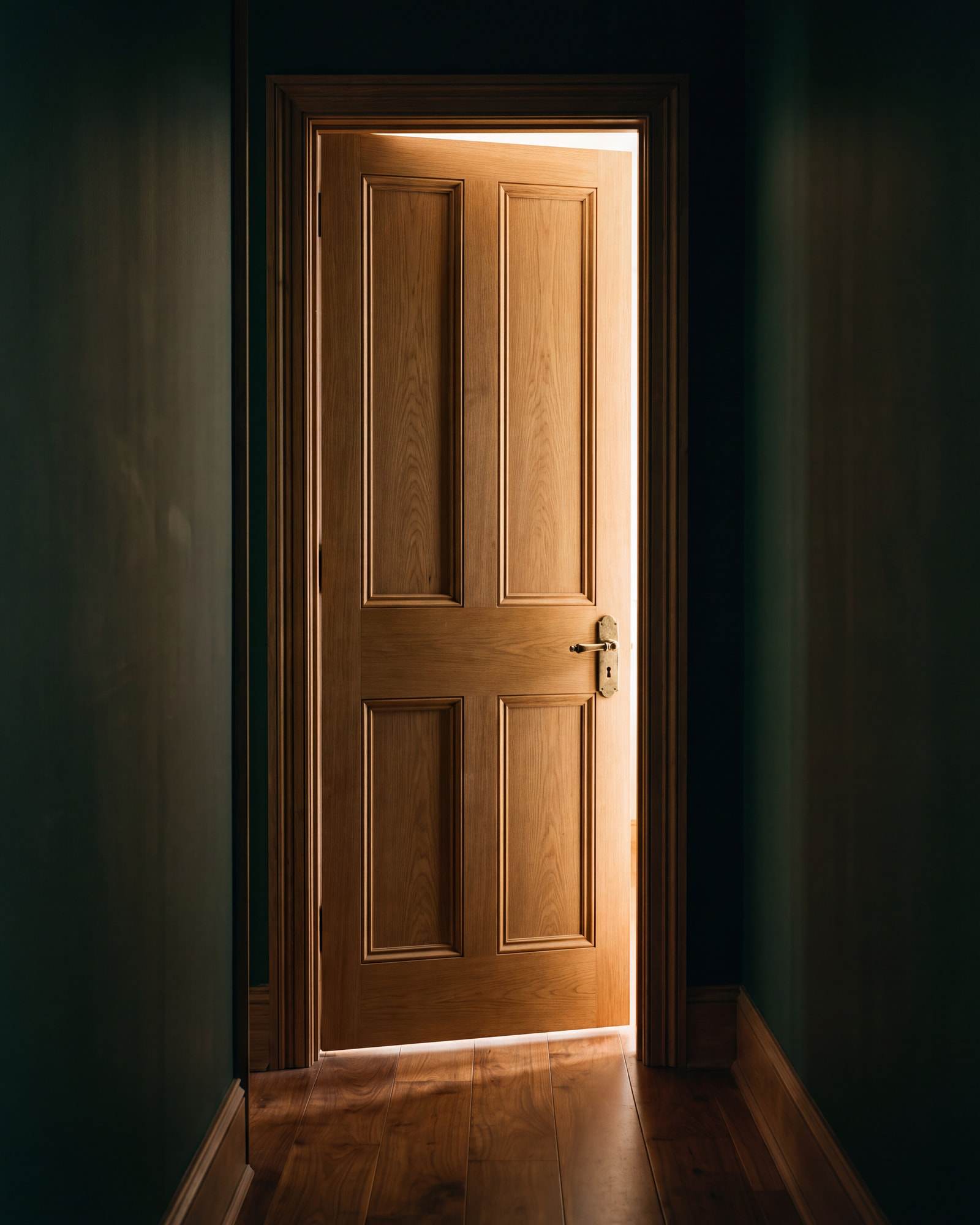 A panelled door standing open at the end of a dark green hallway, warm light beyond it.