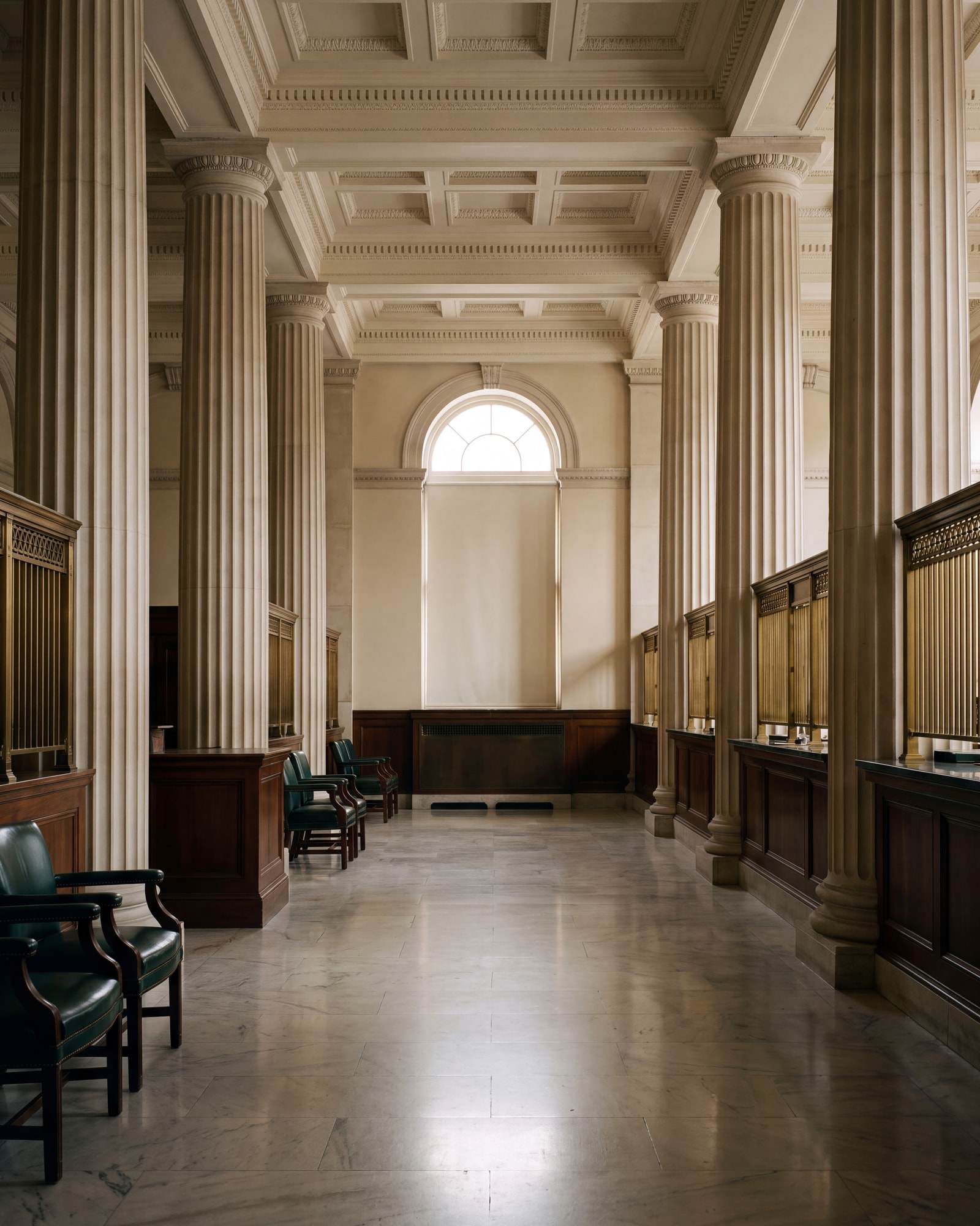 A quiet classical banking hall with limestone columns, brass teller screens, and green leather chairs.