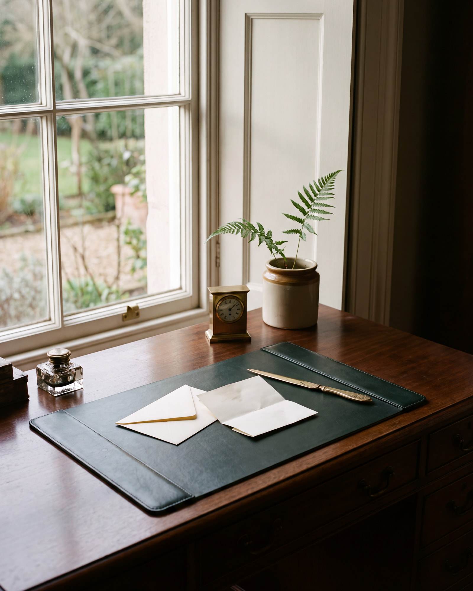A writing desk by a window with an envelope, a letter, and a brass letter opener on a green leather pad.
